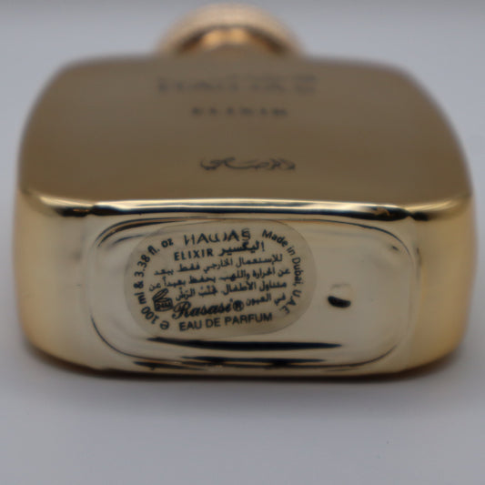 Close-up of the bottom of a gold-toned perfume bottle showing engraved text and a circular Arabic label on a light gray background.