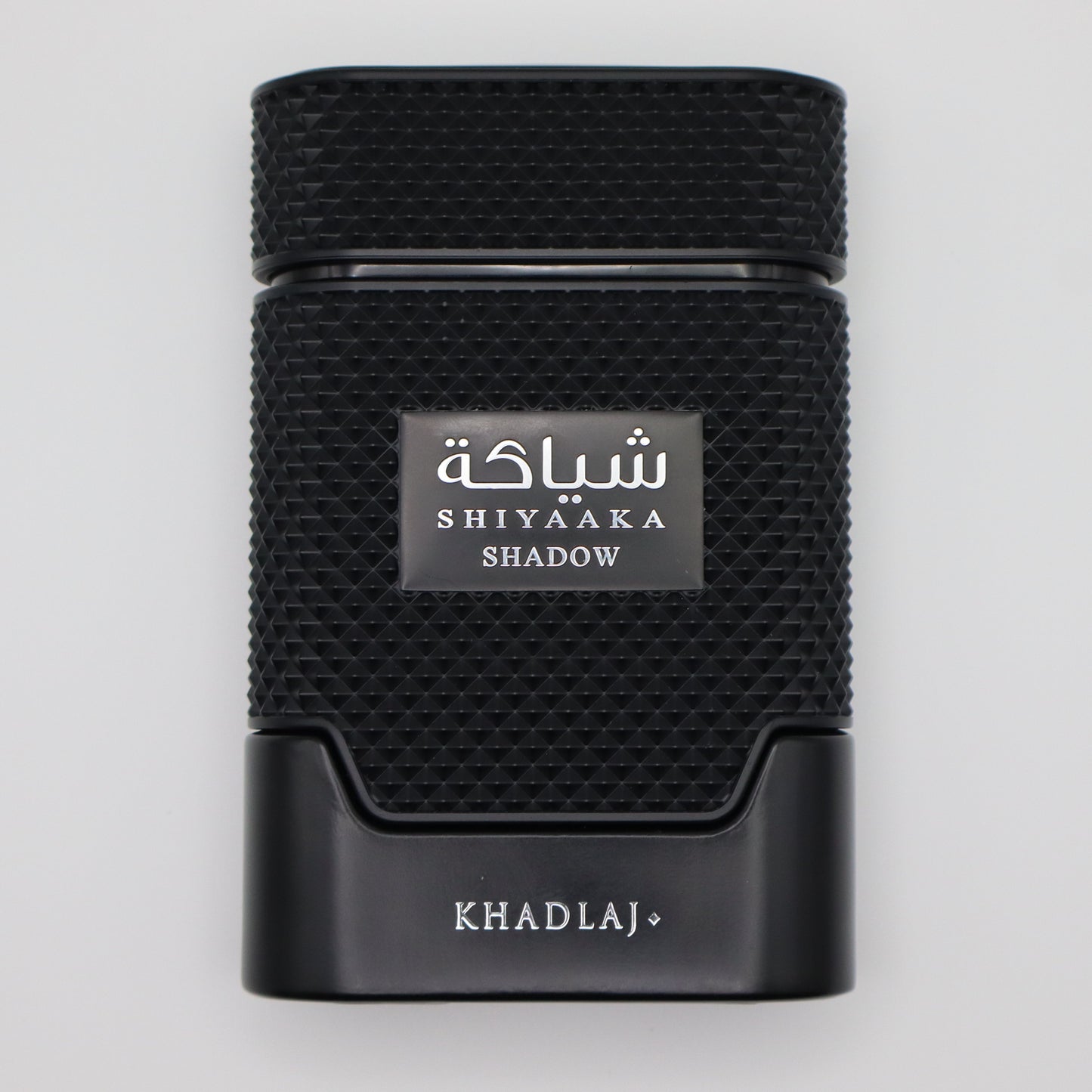 A black textured rectangular perfume bottle with a silver label reading 'SHIYAAKA SHADOW' and 'KHADLAJ+' printed below, centered on a light gray background.
