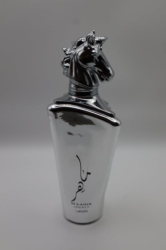 A metallic silver perfume bottle with a sculpted horse head cap and black Arabic calligraphy, labeled 'MAHIR LEGACY' and 'Lattafa', on a neutral gray background.