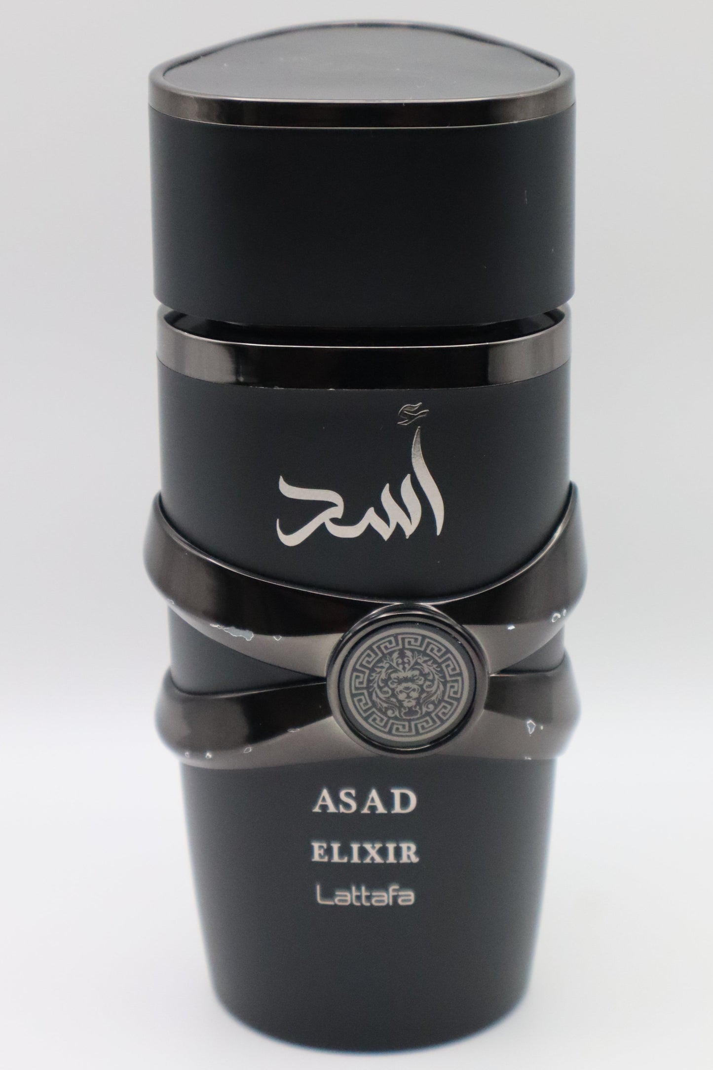 A black cylindrical bottle of Asad Elixir Lattafa perfume with Arabic calligraphy on the front, a silver emblem on the middle band, and white text at the bottom, set against a plain light gray background.