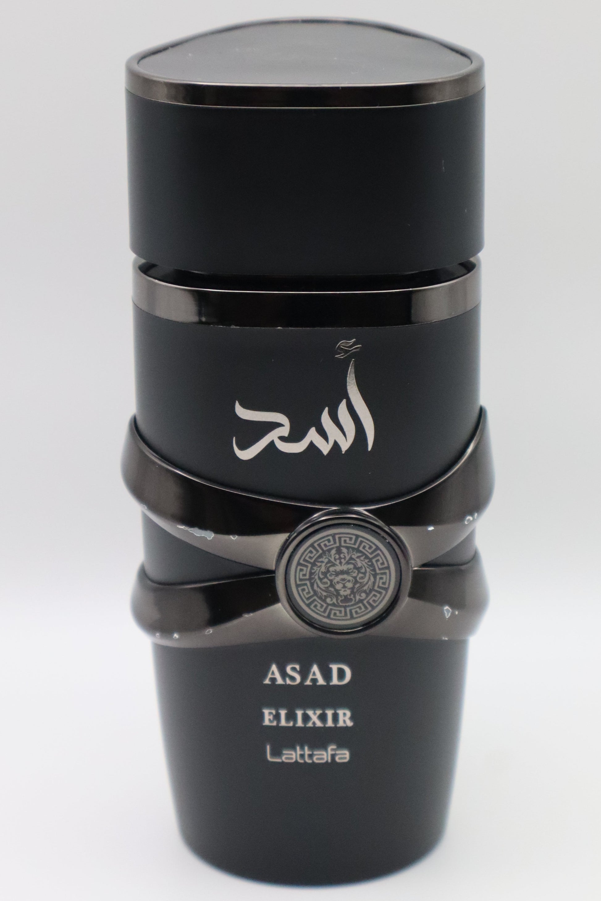 A black cylindrical bottle of Asad Elixir Lattafa perfume with Arabic calligraphy on the front, a silver emblem on the middle band, and white text at the bottom, set against a plain light gray background.