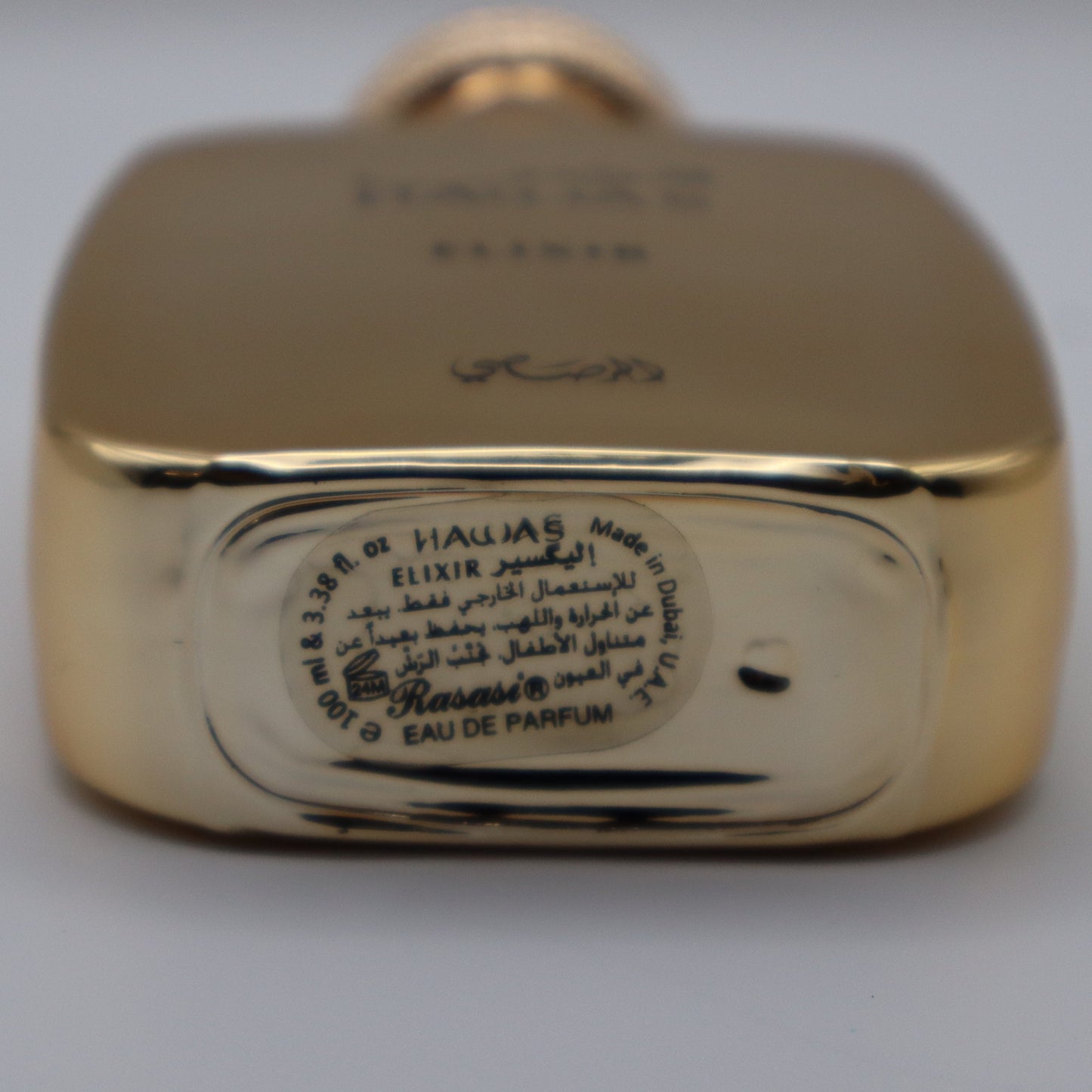 Close-up of the bottom of a gold-toned perfume bottle showing engraved text and a circular Arabic label on a light gray background.