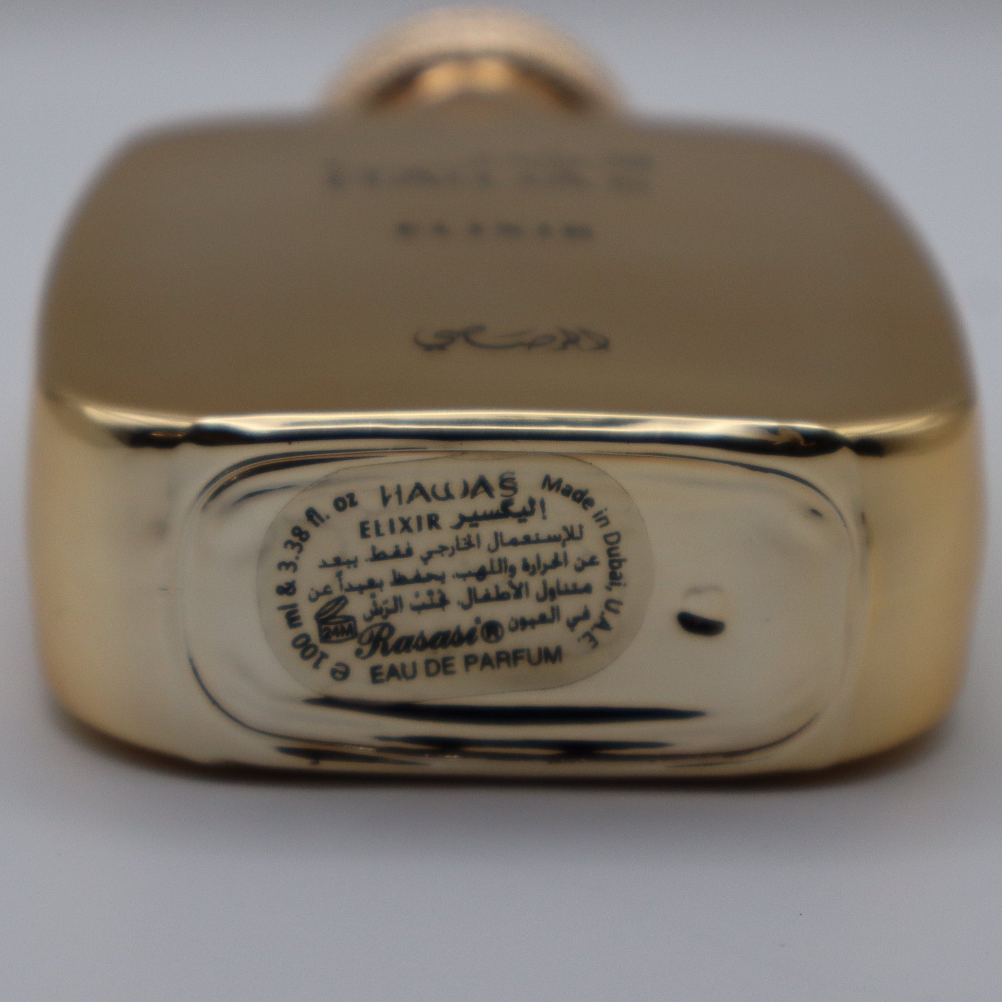 Close-up of the bottom of a gold-toned perfume bottle showing engraved text and a circular Arabic label on a light gray background.