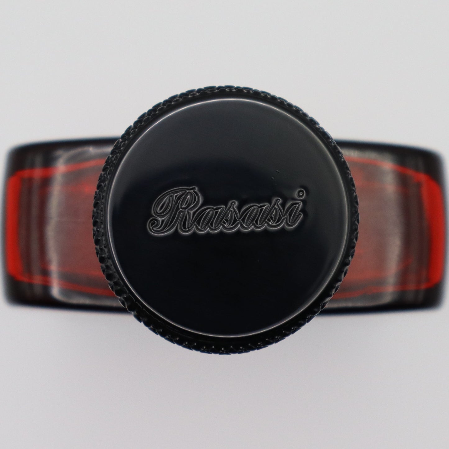 A top-down view of a black circular perfume bottle cap with the brand name 'Rasasi' embossed on top, attached to a black and red perfume bottle.