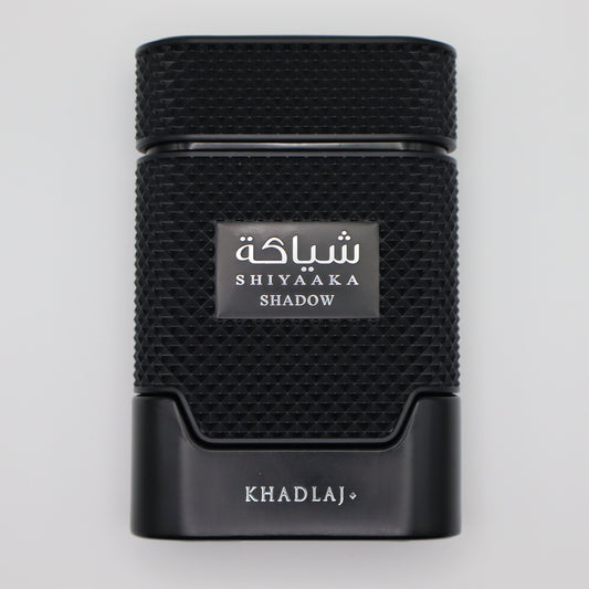 A black textured rectangular perfume bottle with a silver label reading 'SHIYAAKA SHADOW' and 'KHADLAJ+' printed below, centered on a light gray background.