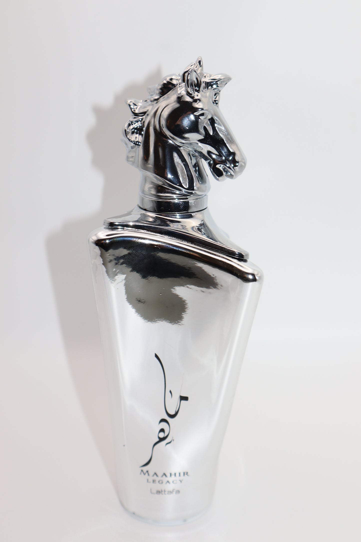 A silver metallic perfume bottle with a sculpted horse head cap and black Arabic calligraphy, labeled 'MAAHIR LEGACY' and 'LATTAFA' on a plain white background.