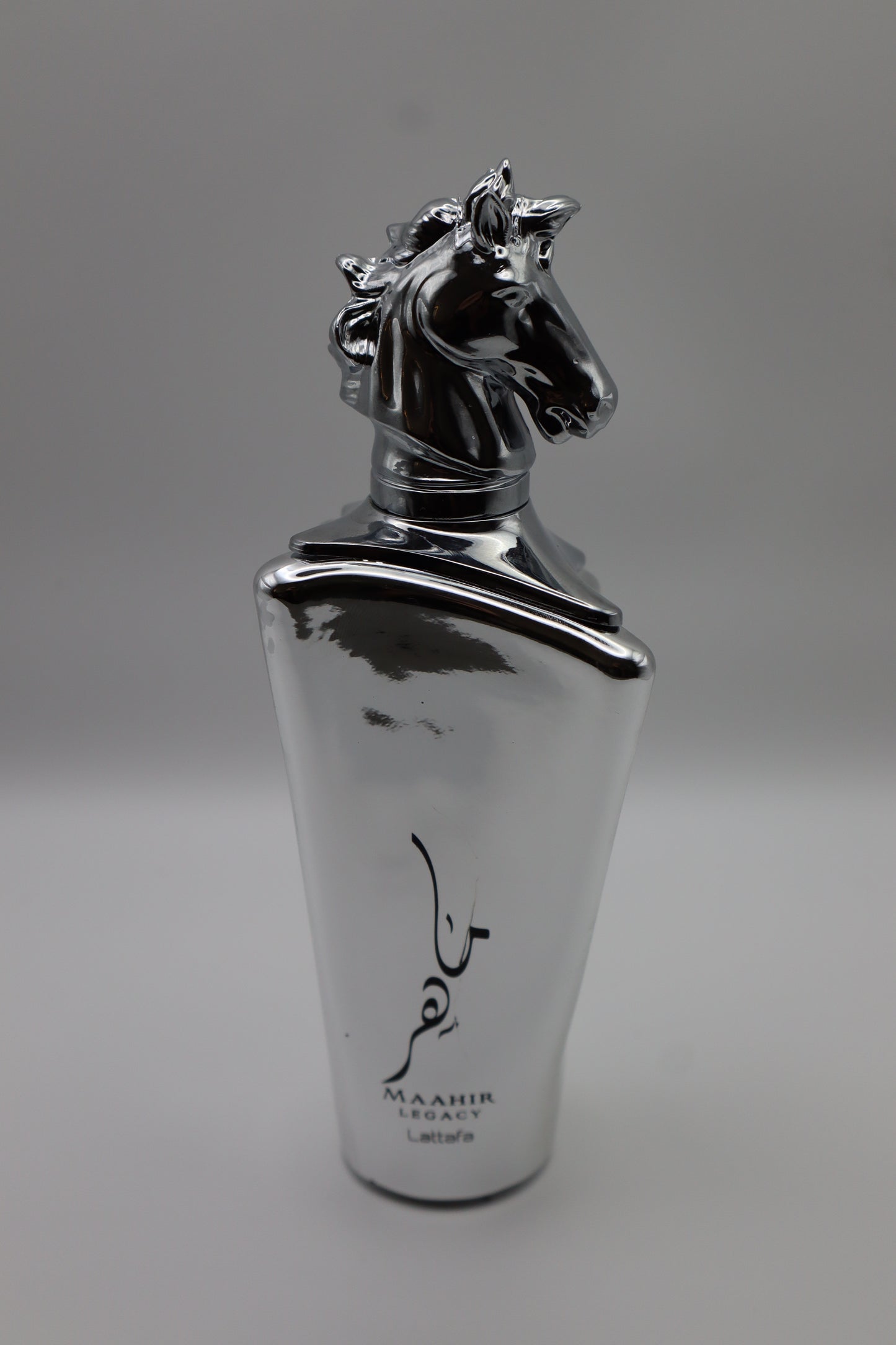 A metallic silver perfume bottle with a sculpted horse head cap and black Arabic calligraphy, labeled 'MAHIR LEGACY' and 'Lattafa', on a neutral gray background.