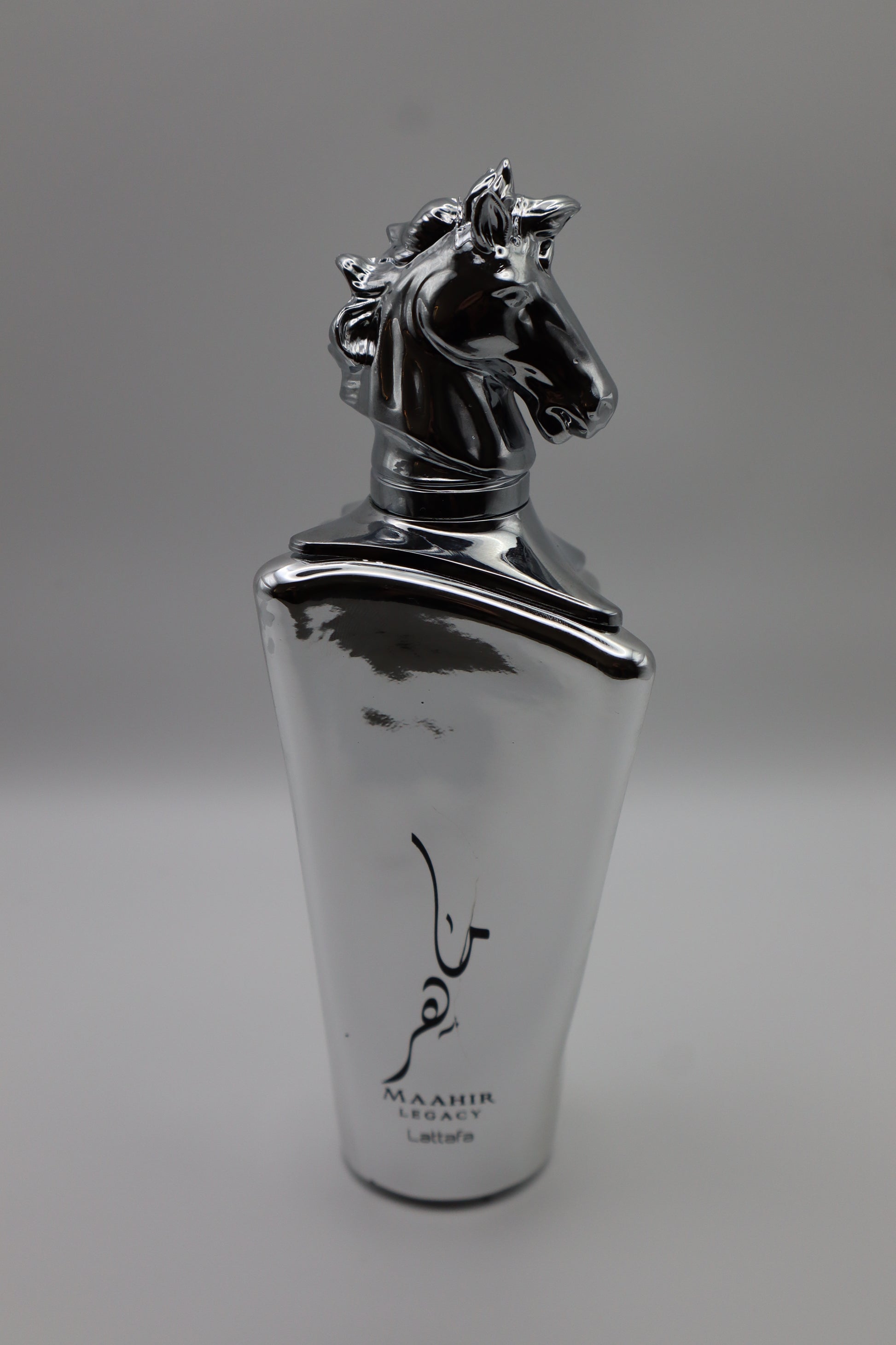 A metallic silver perfume bottle with a sculpted horse head cap and black Arabic calligraphy, labeled 'MAHIR LEGACY' and 'Lattafa', on a neutral gray background.