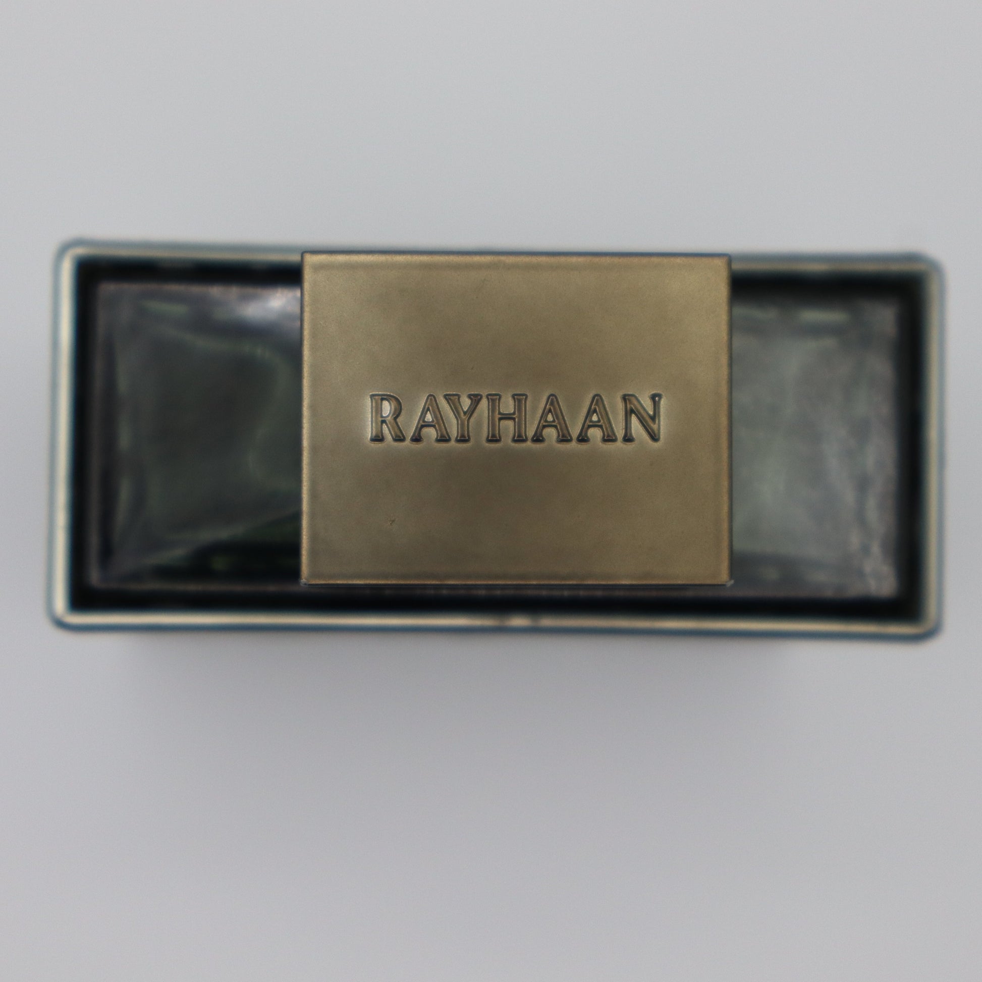 A rectangular metallic perfume bottle with a central brass-colored cover engraved with the brand name 'RAYHAAN', set against a plain light gray background.